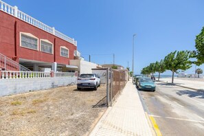 Une rue côtière bordée de voitures garées, avec un trottoir et un immeuble à la façade rouge et au balcon blanc.