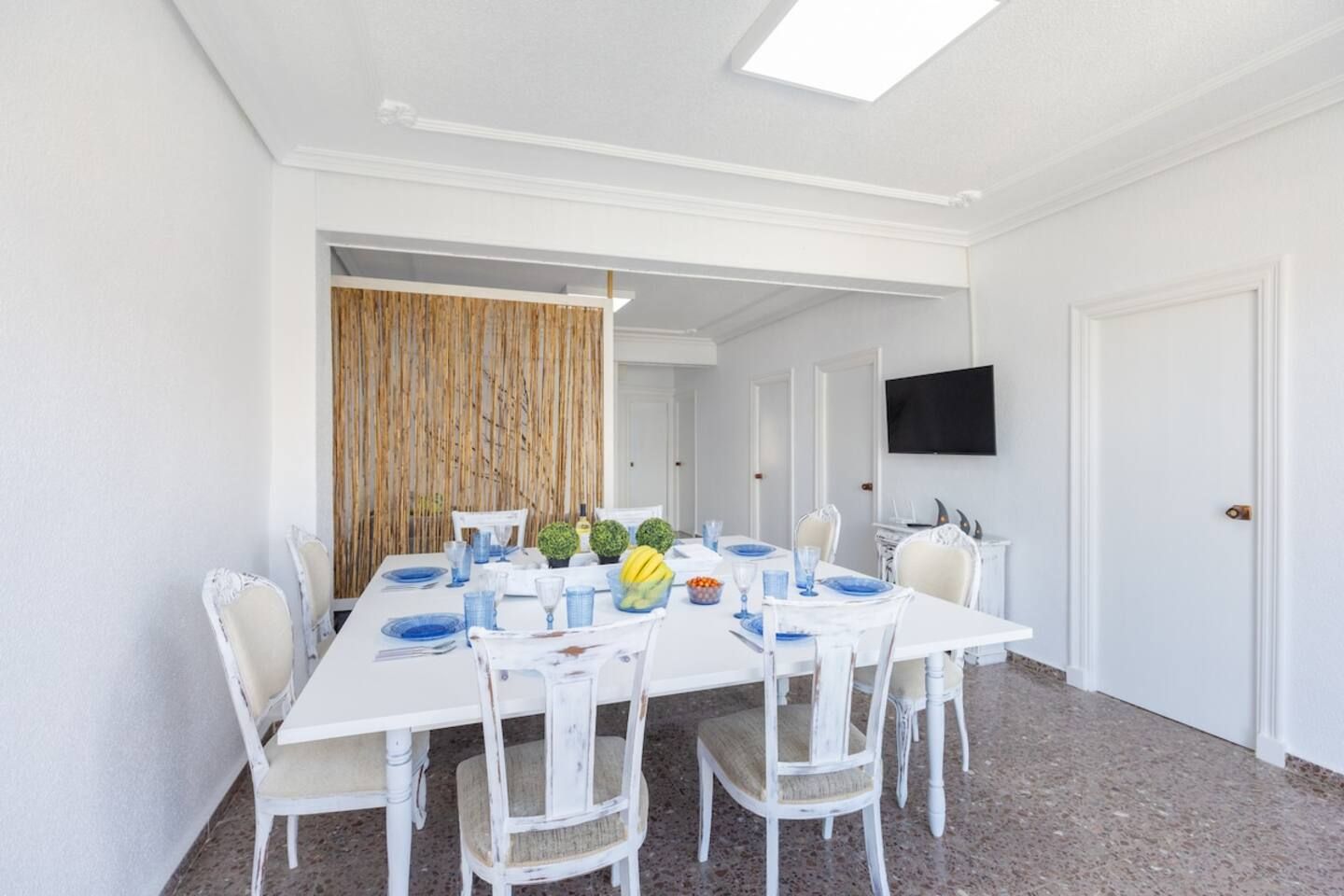 A dining area with a white table and chairs, a television mounted on the wall, and a door to the right.