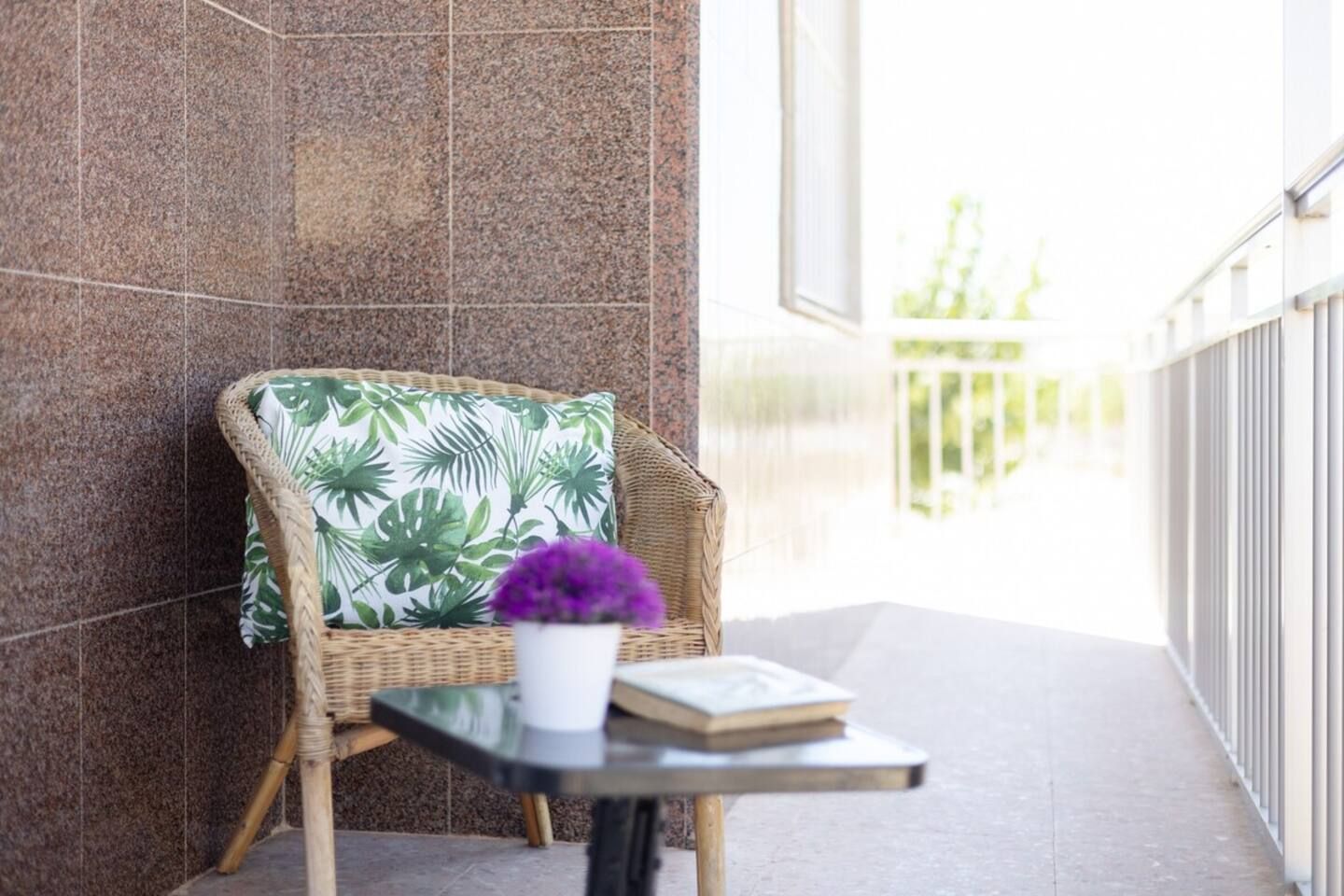 Una silla de mimbre con cojín de hoja tropical, una mesita con un libro y una planta en maceta, y vista a un balcón con barandilla.