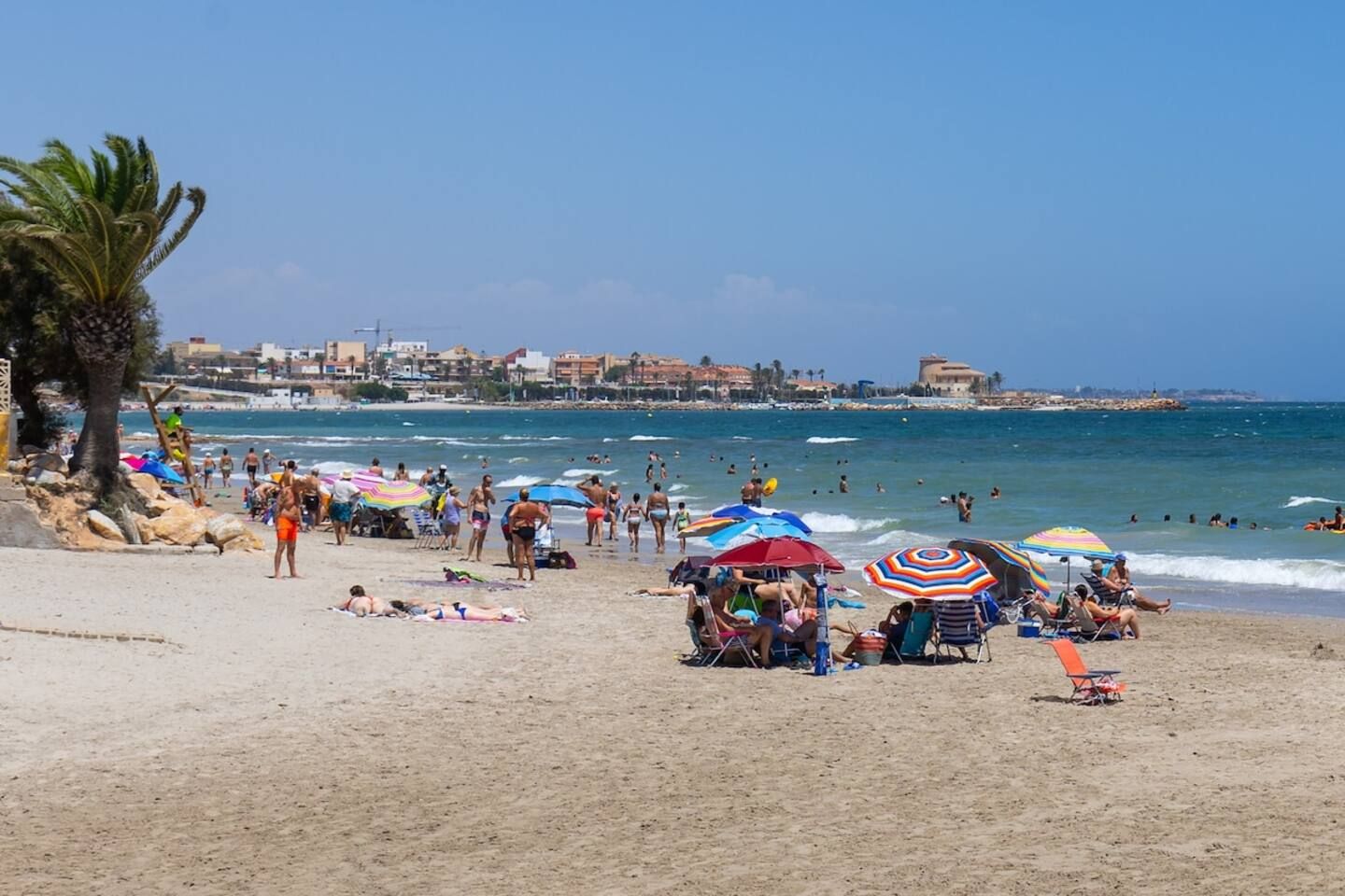 A beach with people sunbathing and swimming, umbrellas, and a clear sky.