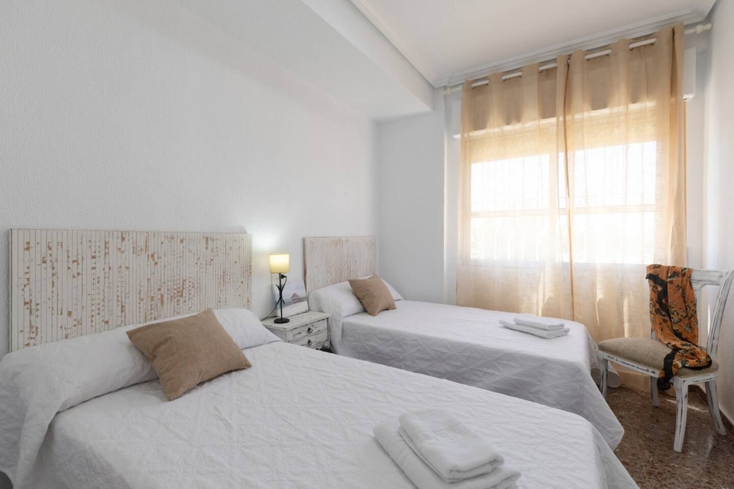 A hotel room with two beds, a nightstand with a lamp, a chair, and a window with curtains.