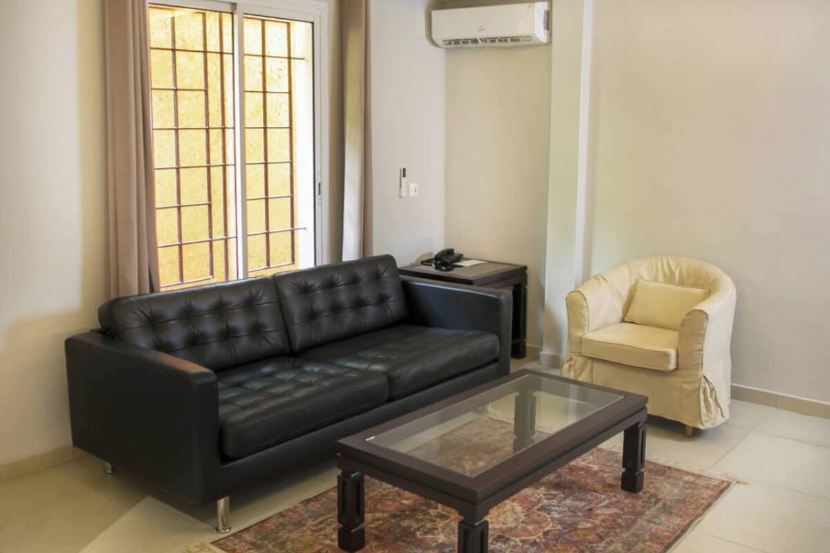 A living room with a black sofa, a glass coffee table, a beige armchair, a small side table with a lamp, and a window with curtains.