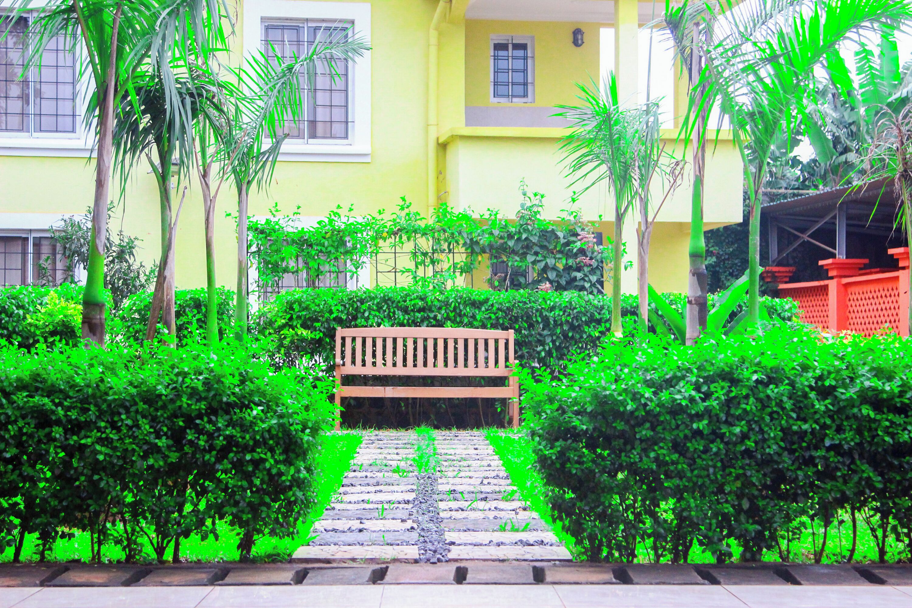 A pathway leading to a wooden bench surrounded by lush green hedges and palm trees, with a yellow building in the background.