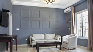 Un salón con pared gris, un sofá blanco, una mesa de centro de madera, un sillón blanco, un televisor sobre un mueble bajo y una ventana con cortinas.