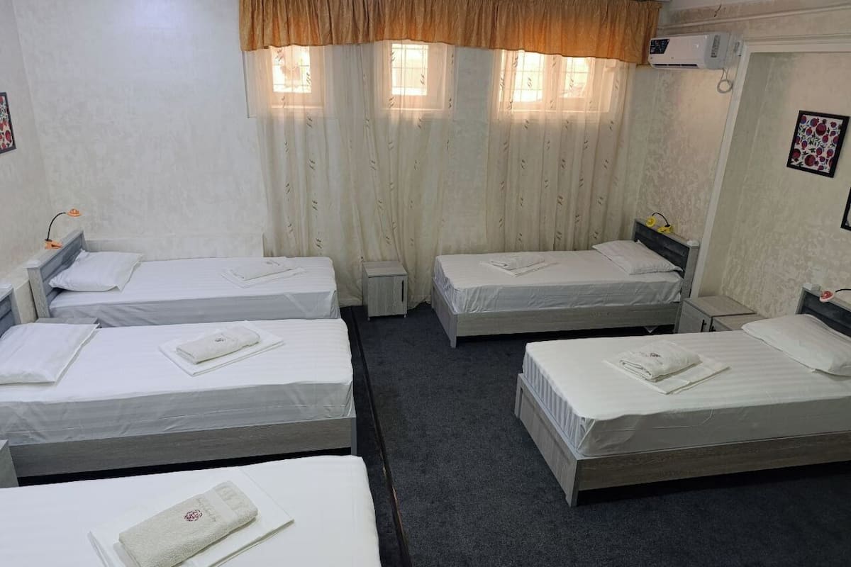 Ein Hotelzimmer mit vier Einzelbetten, einem kleinen Nachttisch, einem Fenster mit Vorhängen und einer Klimaanlage.