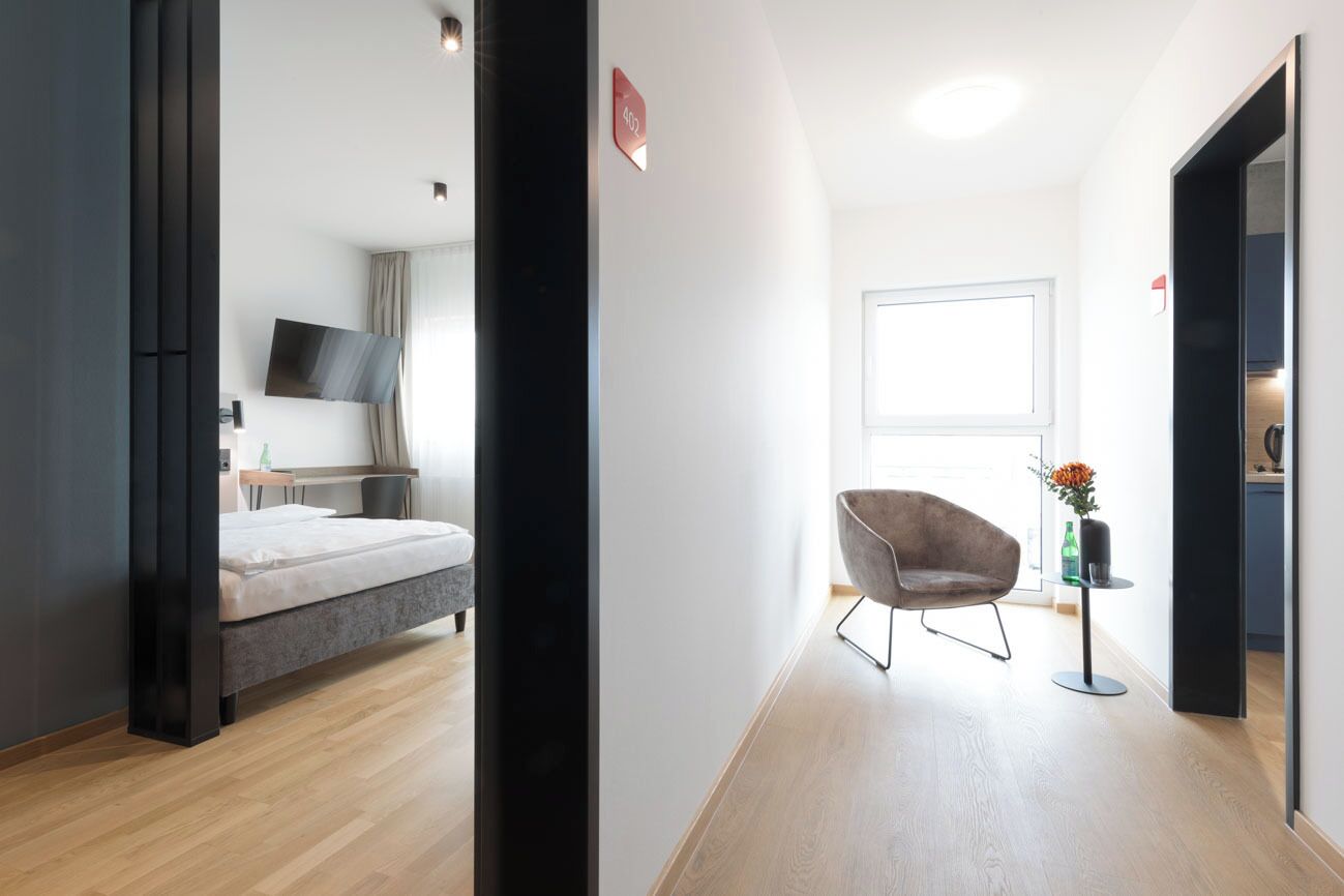 Ein modernes Hotelzimmer mit Bett, Flachbildfernseher, Sessel und einem kleinen Tisch mit Blumenvase.