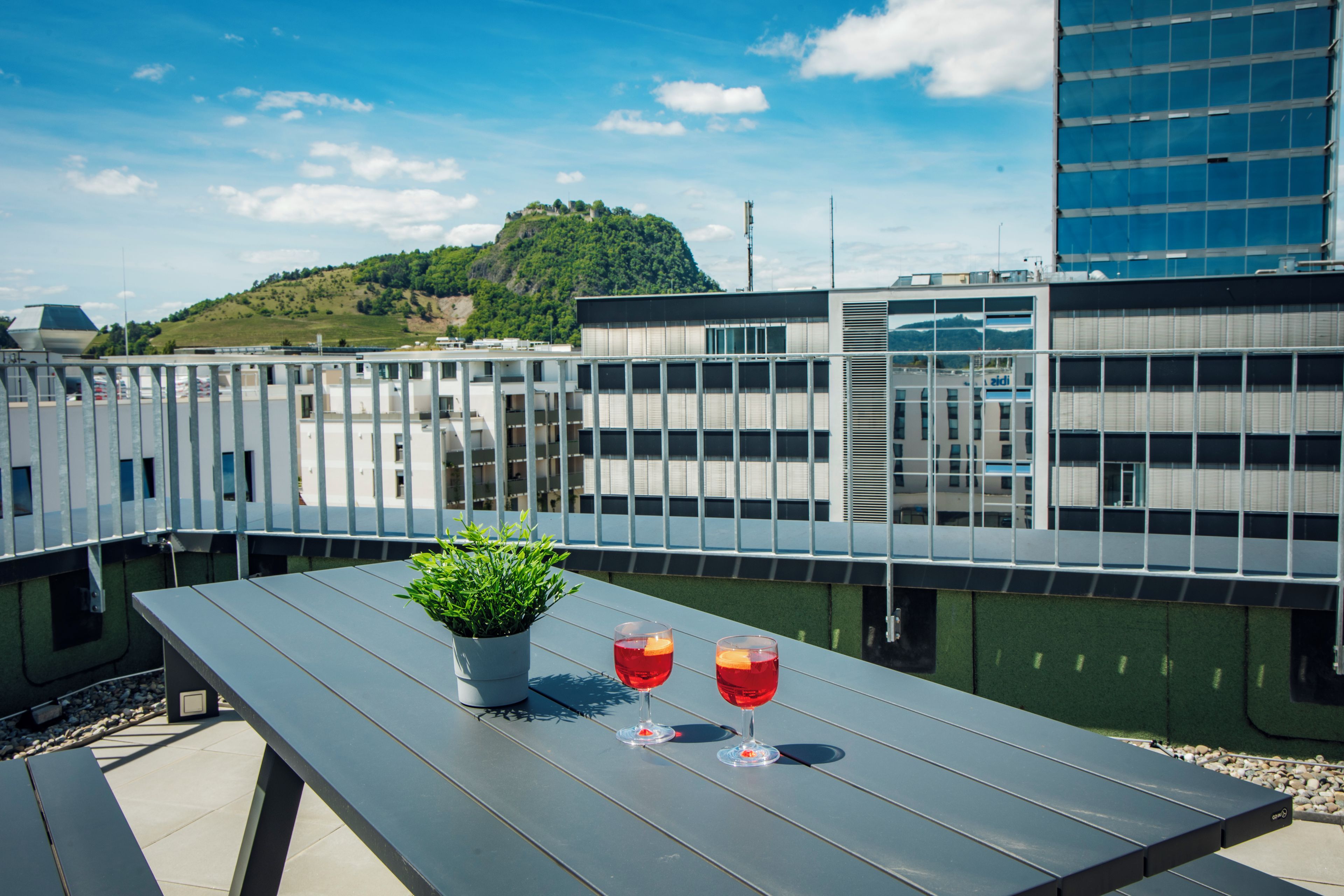 A rooftop terrace with a table, two glasses of red beverage, and a potted plant.