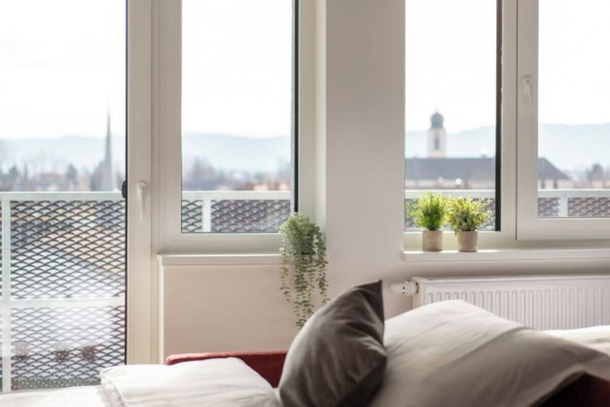 A room with a bed, a window with a view of a cityscape, and a plant on the windowsill.