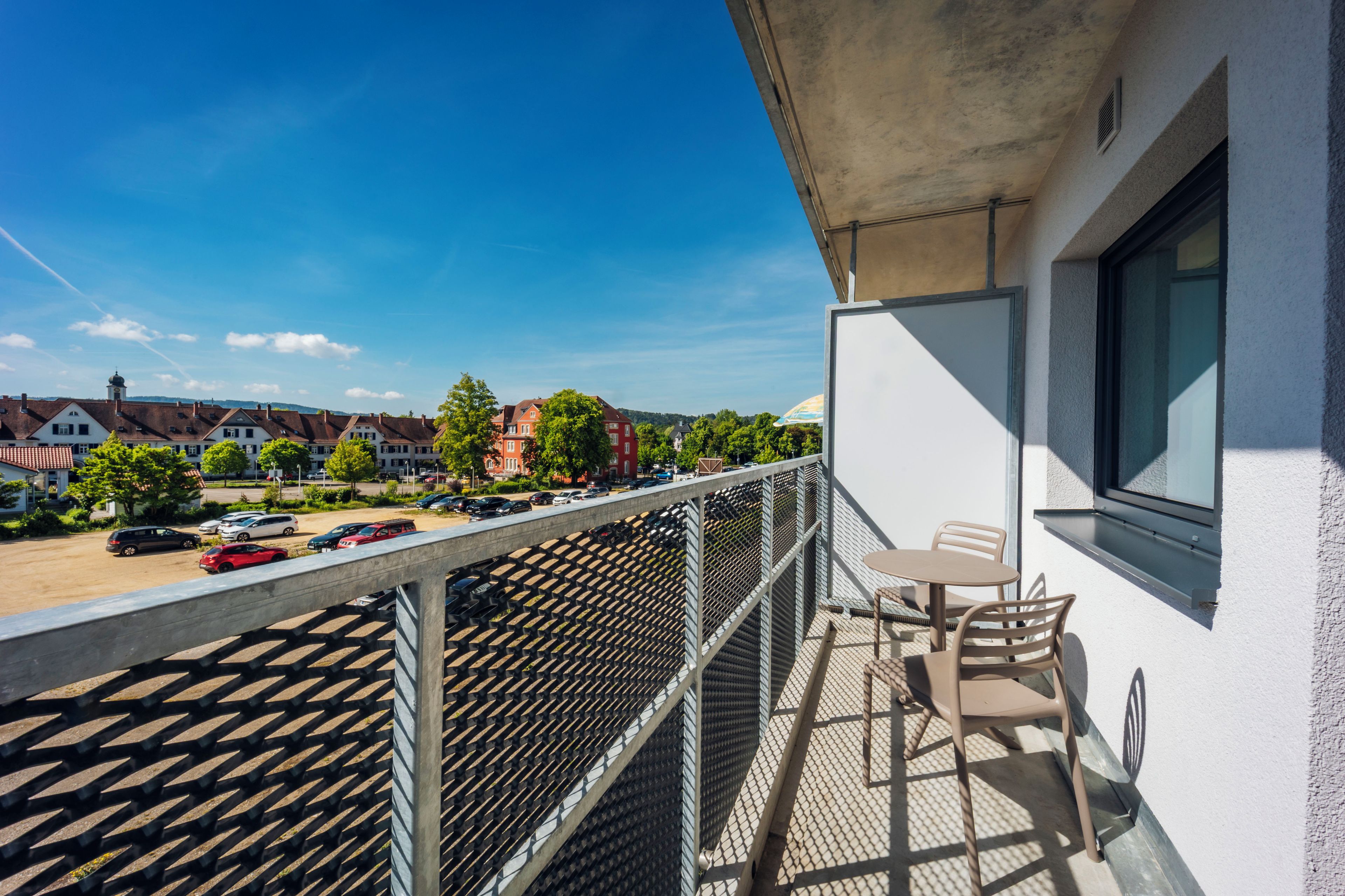 A balcony with a table and chairs, overlooking a parking area and buildings.