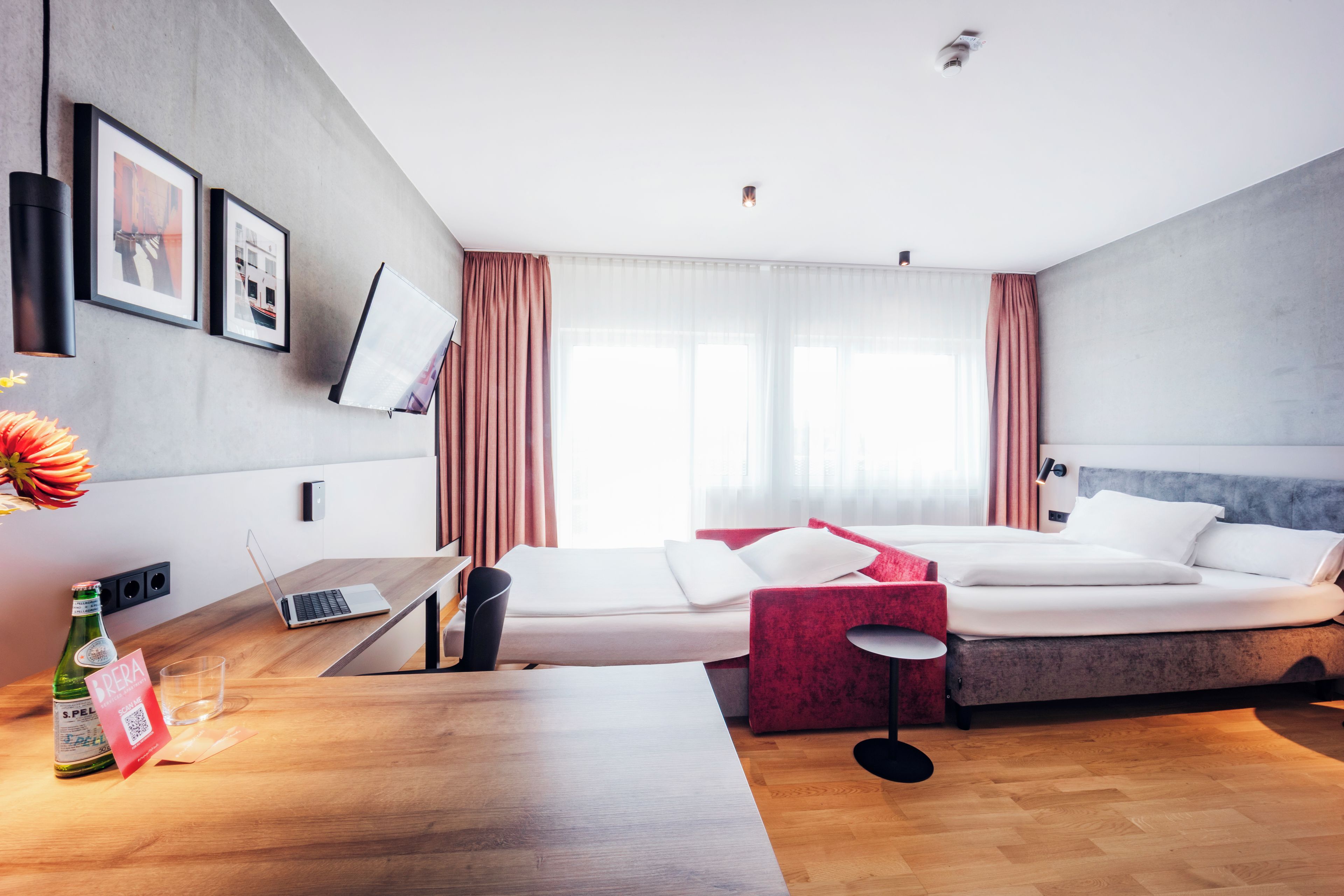A modern hotel room with a wooden desk, a laptop, a bottle of wine, and a red chair.
