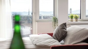 A room with a bed, a window with a view of a cityscape, and a plant on the windowsill.