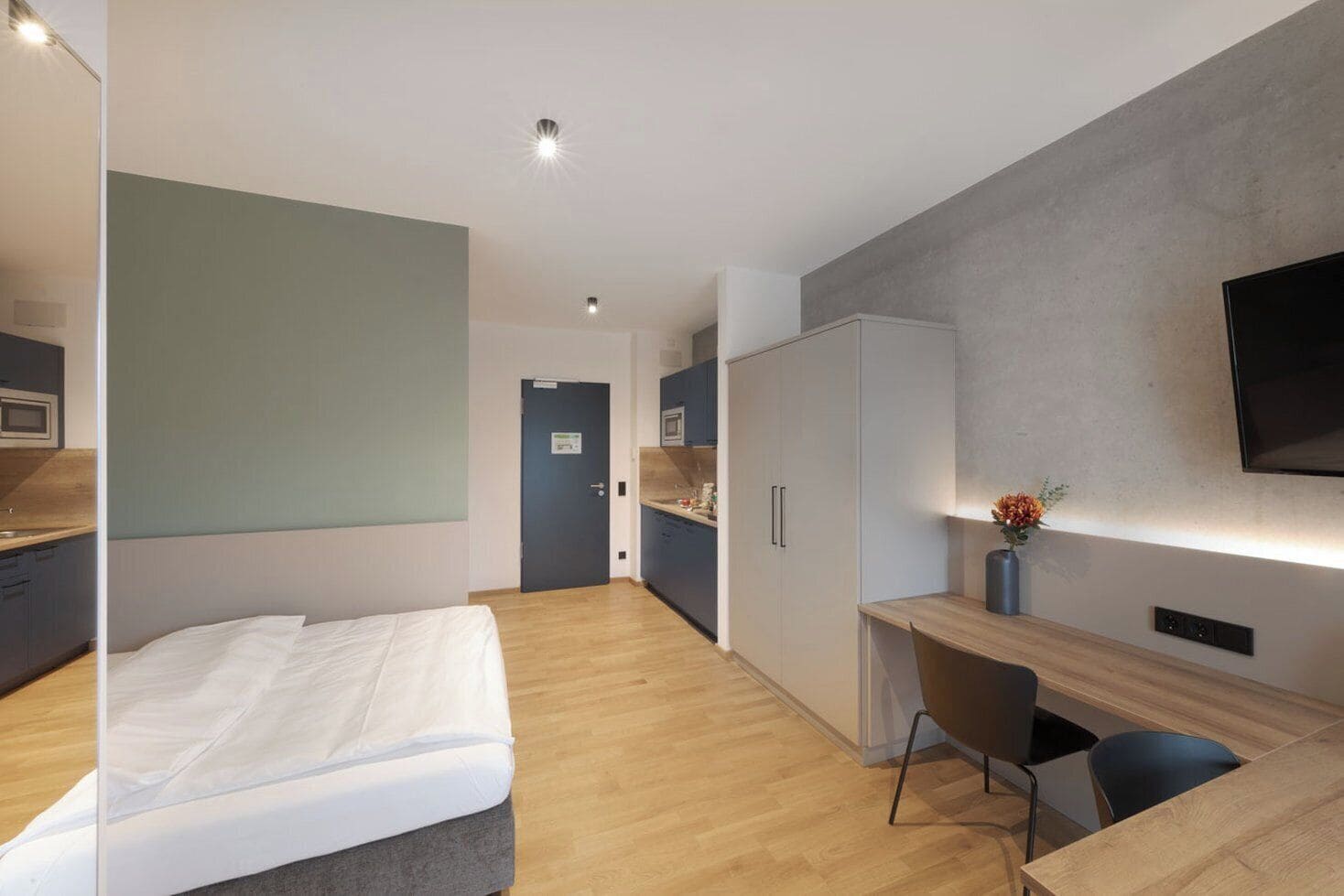 A modern hotel room with a bed, a desk, a chair, a TV, and a kitchenette.