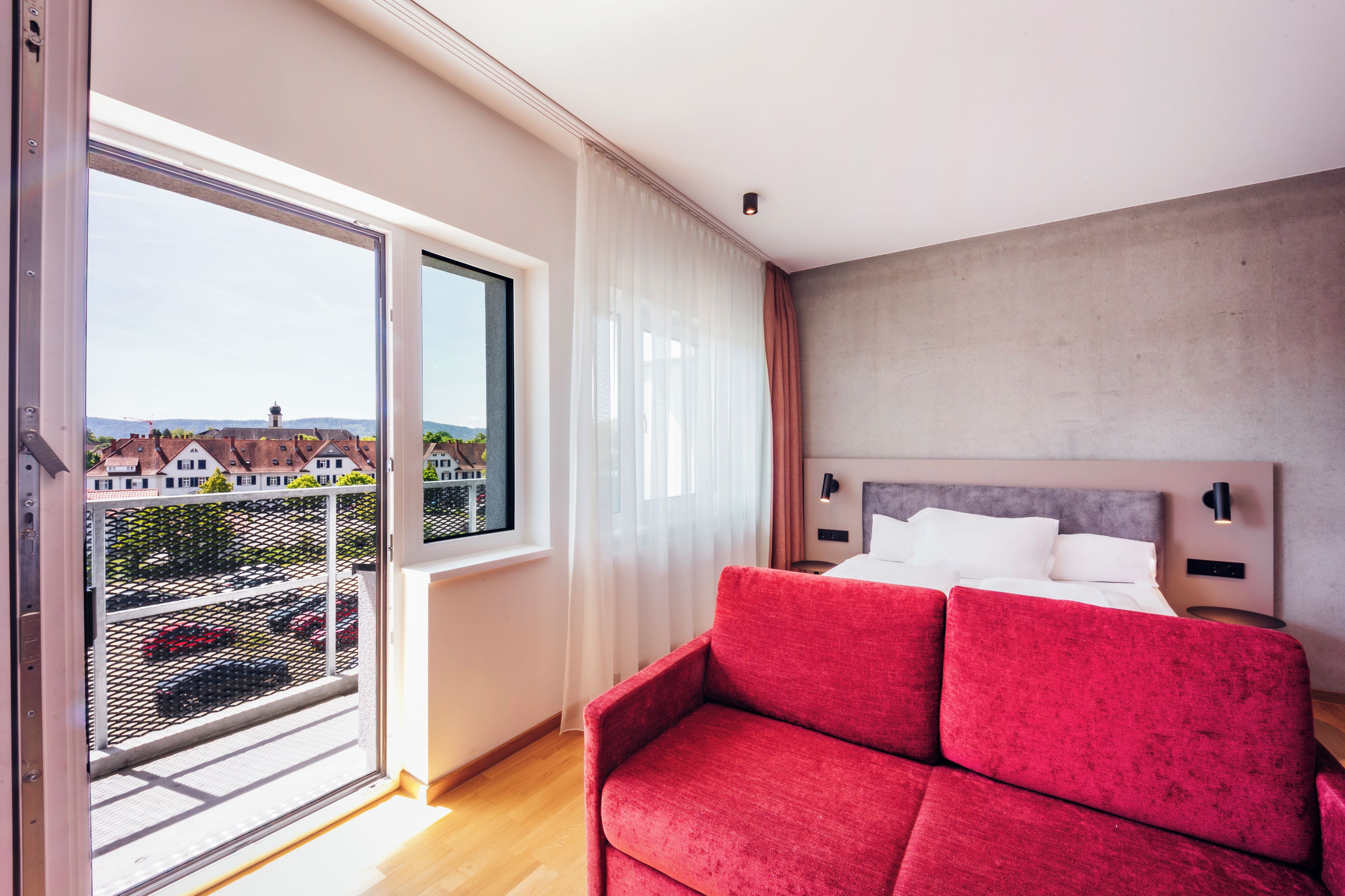 A hotel room with a balcony, a bed, a red sofa, and a view of buildings and a crane.