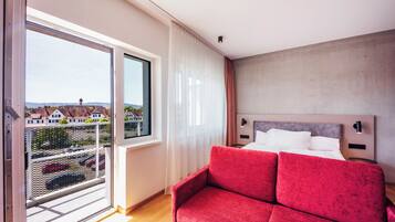 A hotel room with a balcony, a bed, a red sofa, and a view of buildings and a crane.
