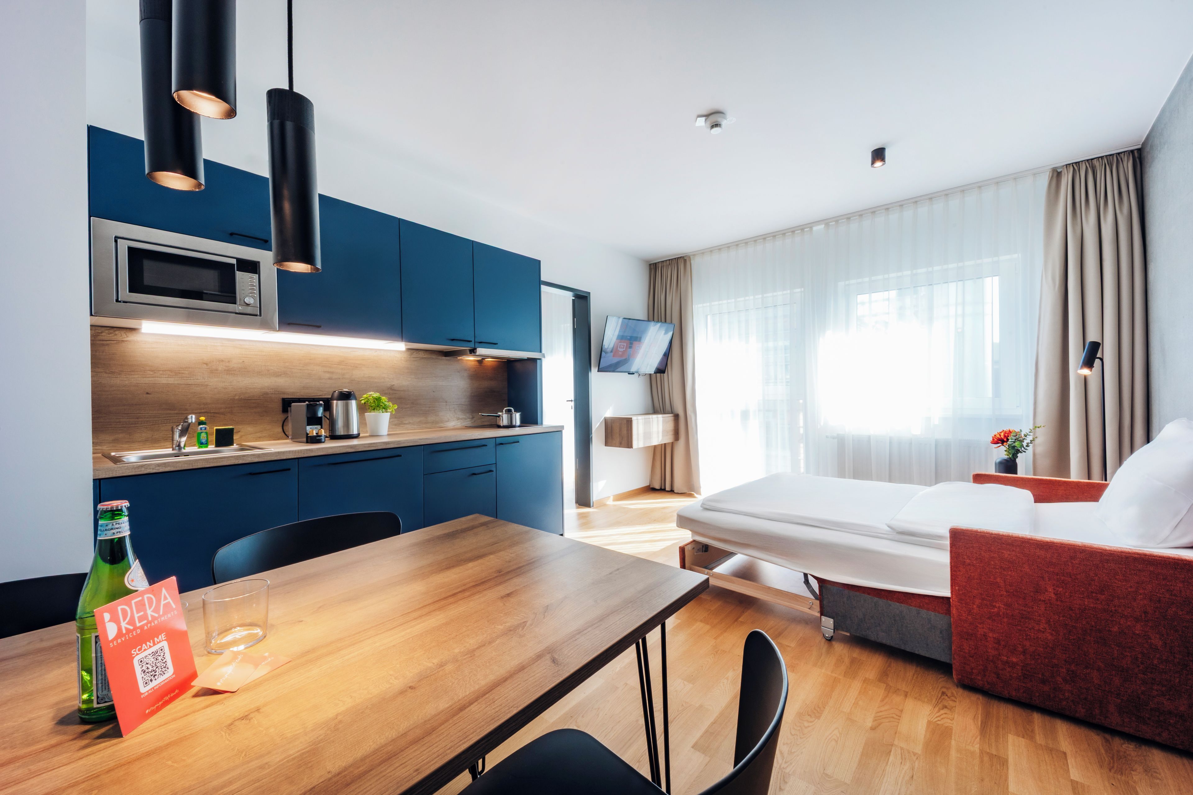 A modern hotel room with a wooden dining table, a sofa, a bed, a TV, and a kitchen area with blue cabinets and a microwave.