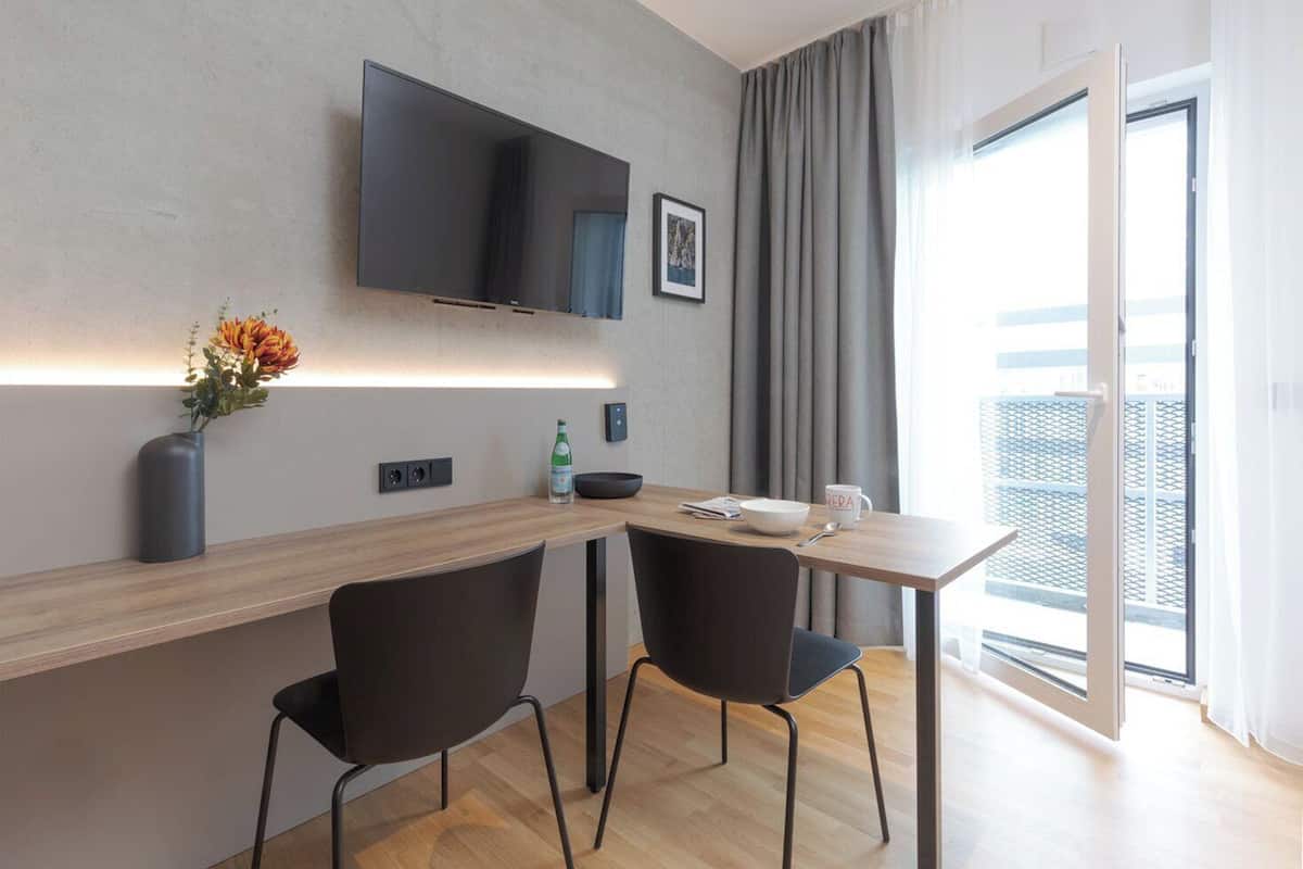 Une chambre d’hôtel moderne avec un bureau en bois, deux chaises noires, un téléviseur à écran plat fixé au mur, un petit vase avec des fleurs, une bouteille d’eau, un bol, une cuillère et une tasse sur la table.