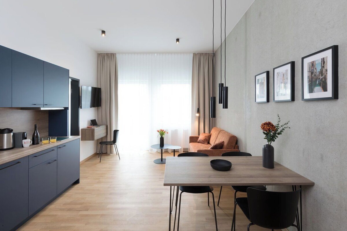 A modern kitchen with dark cabinets, a dining area with a wooden table and chairs, and a living space with a sofa and a coffee table.