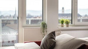 A room with a bed, a window with a view of mountains and a lighthouse, and two potted plants on the windowsill.