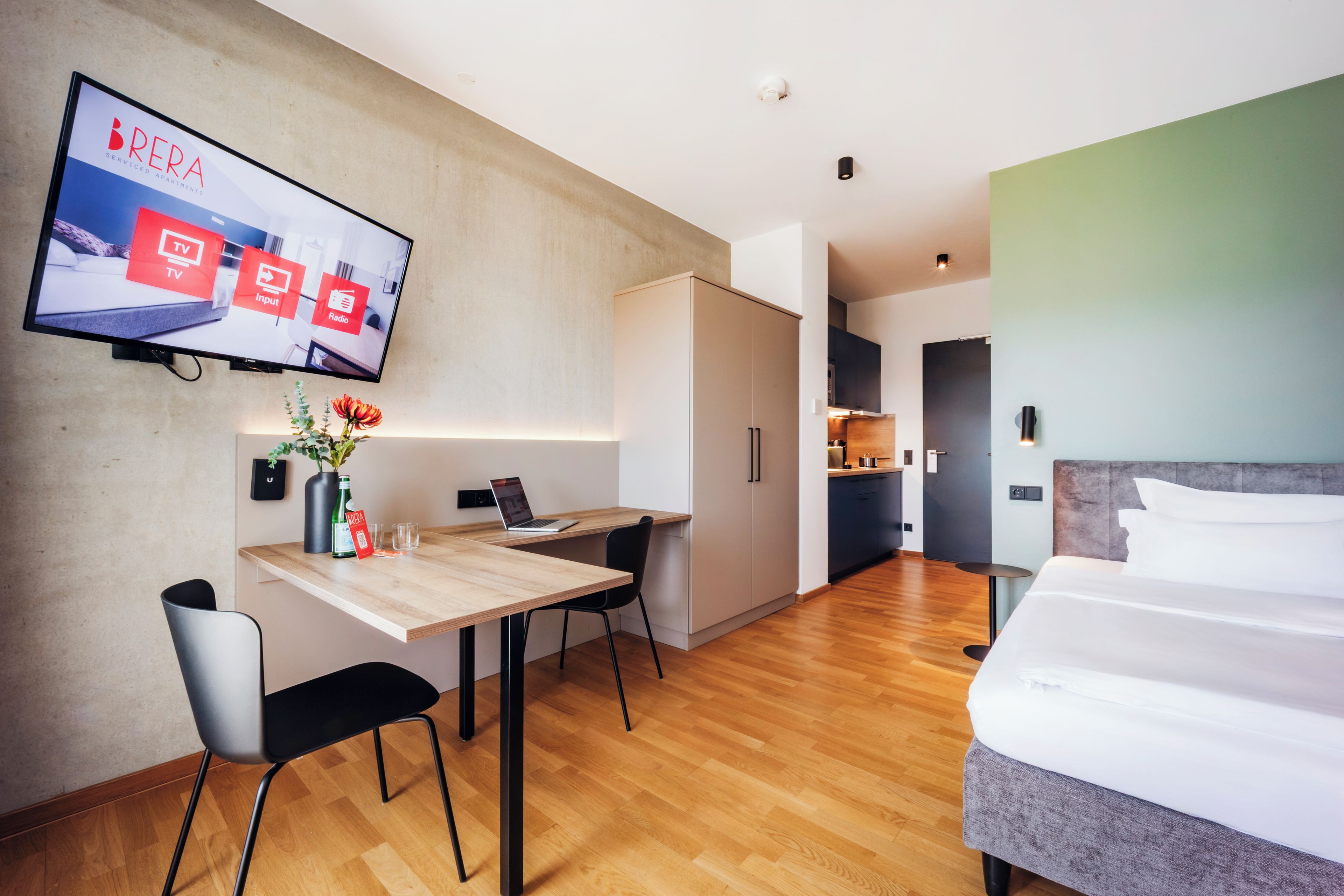 A modern hotel room with a wooden desk, a bed, and a TV mounted on the wall displaying a service interface.