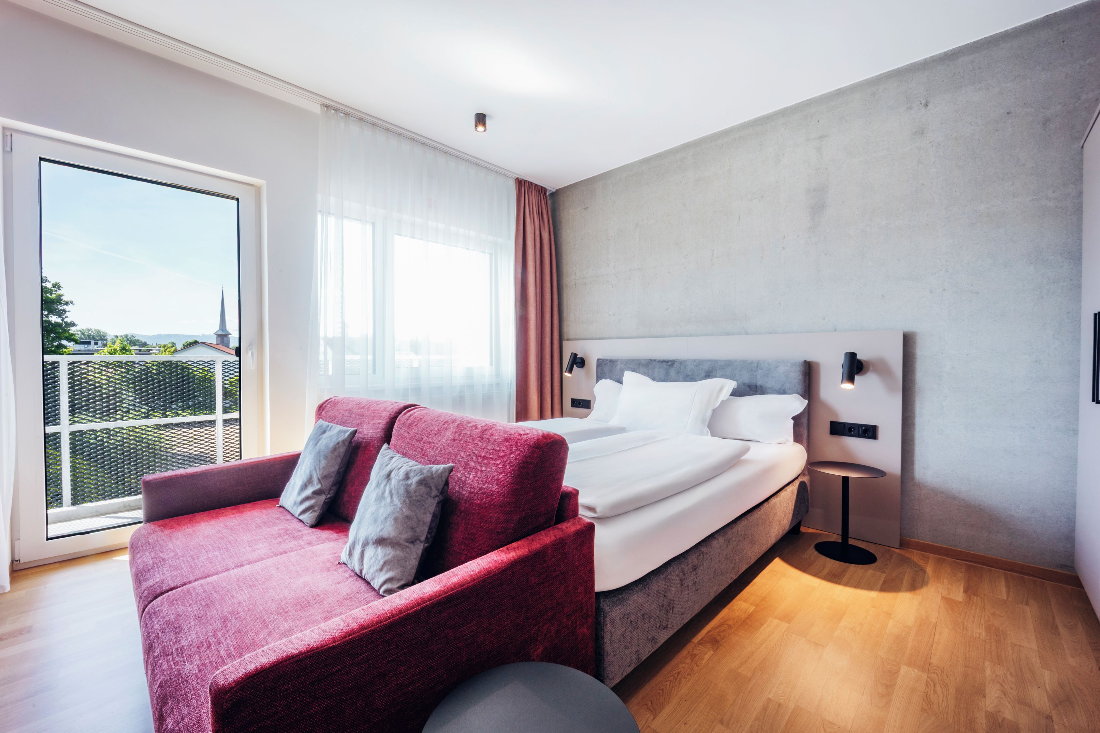 A modern hotel room with a large bed, a red sofa, and a view of the outdoors.