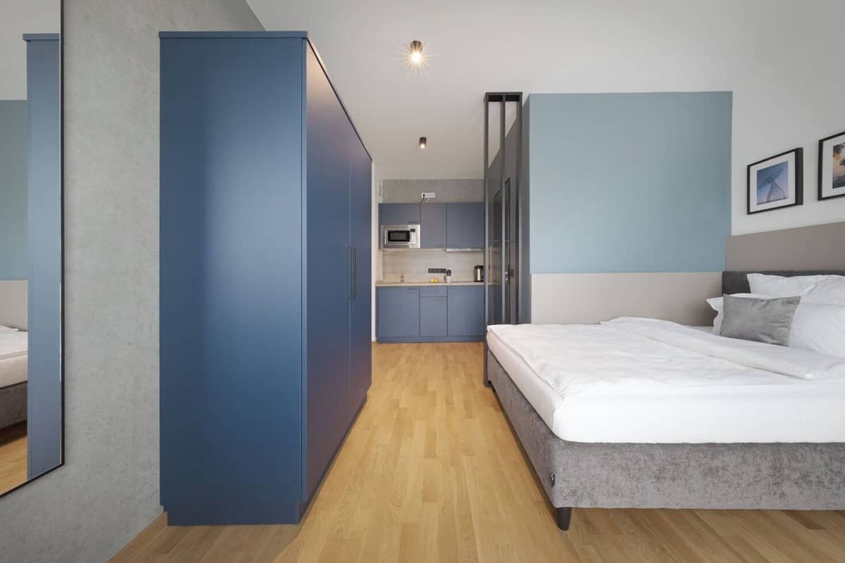 Une chambre moderne avec un grand lit, une armoire intégrée et un coin cuisine à l’arrière-plan.
