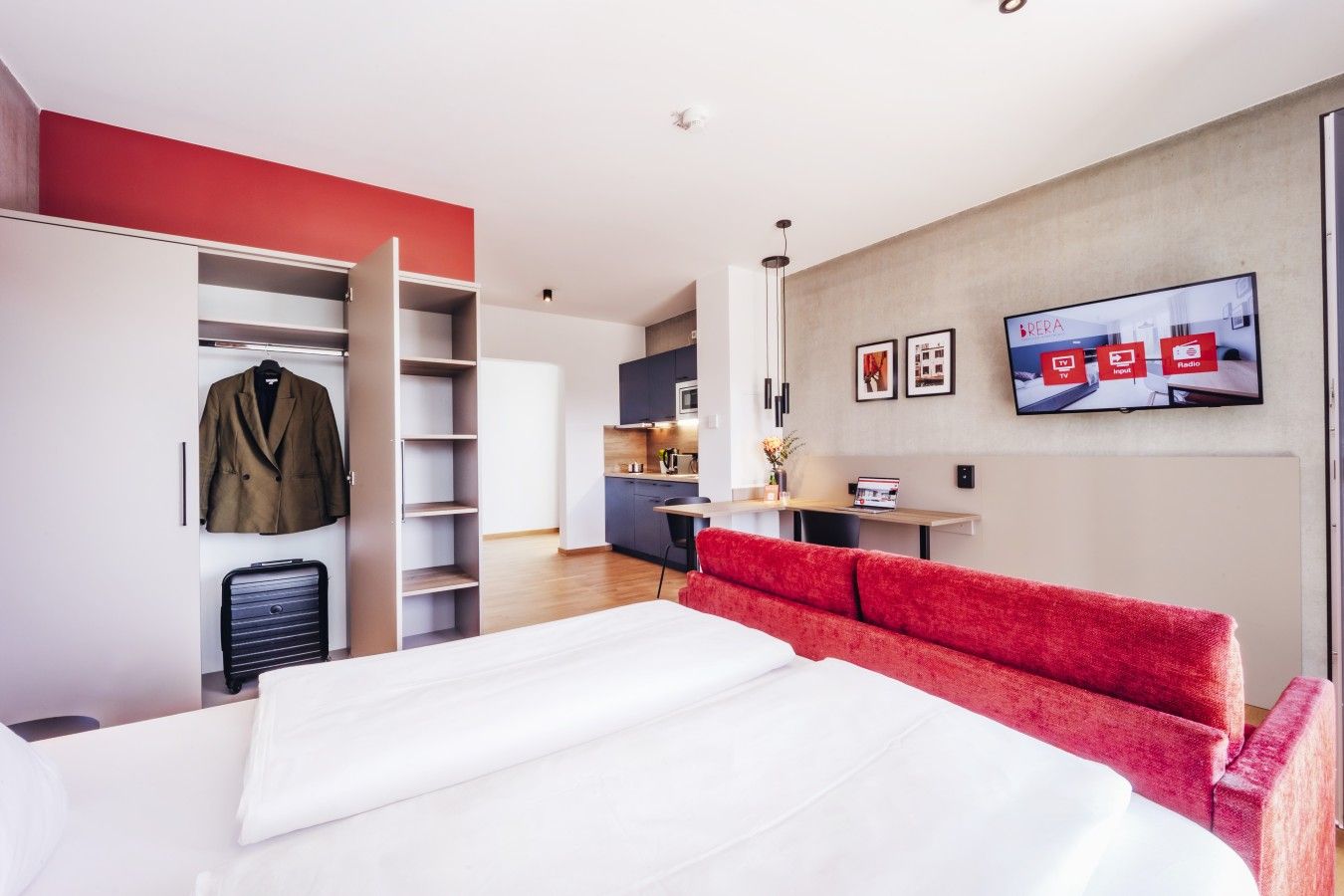 Una camera d'albergo con un letto, un divano rosso, un armadio, una scrivania e un televisore che mostra un menù.