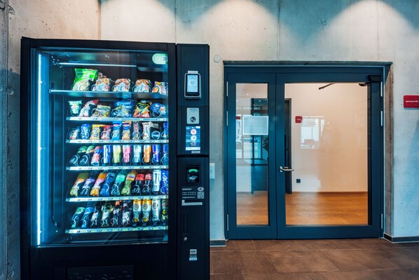 Máquina expendedora llena de bocadillos y bebidas, una puerta con panel de vidrio y un cartel que indica 'Apartments 001-004'.