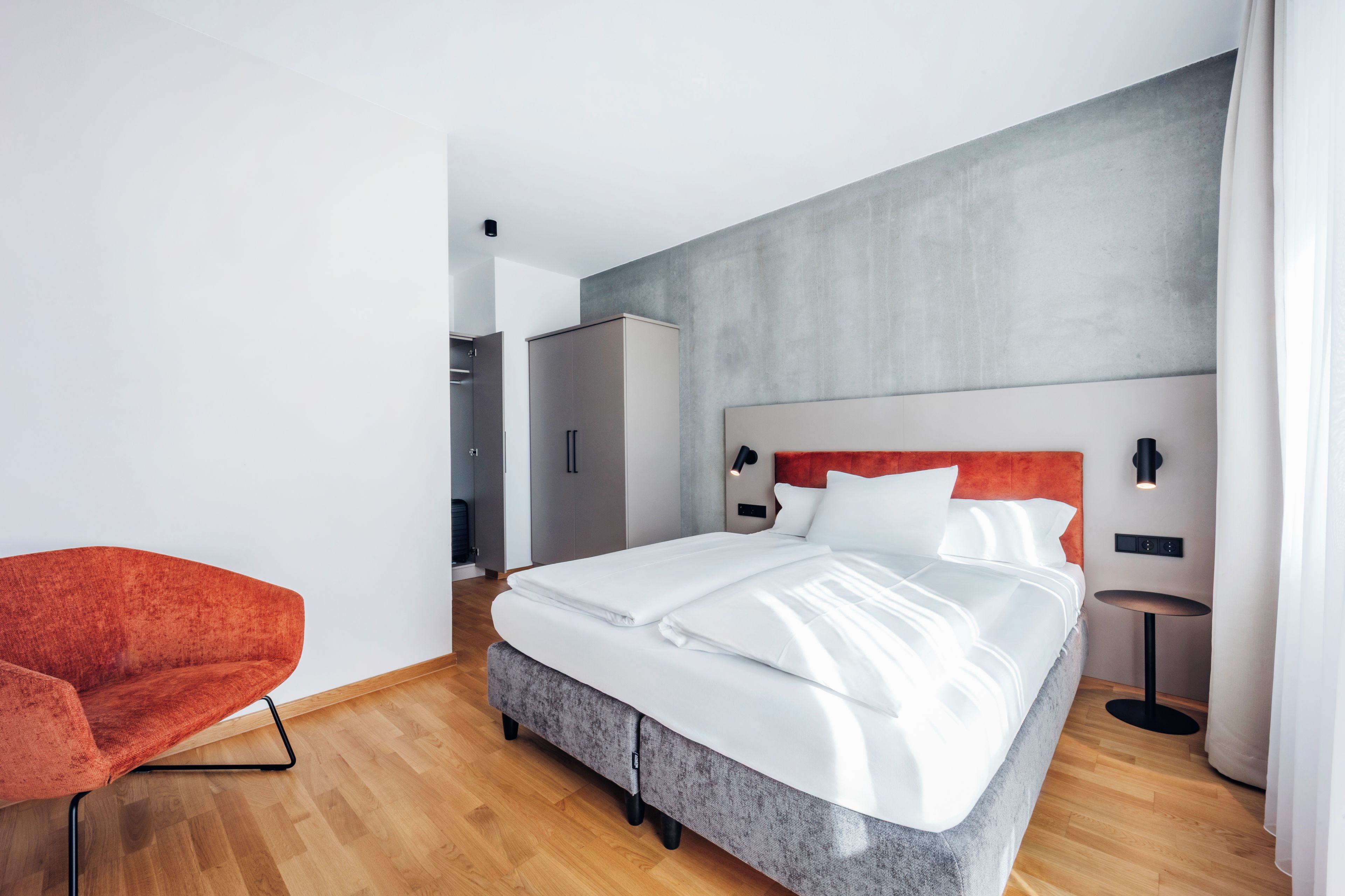 A modern hotel room with a large bed, a red chair, a wardrobe, and a small table.