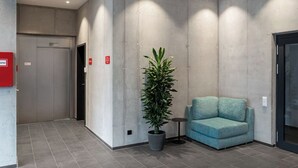 A modern lobby with a concrete wall, a potted plant, a blue sofa, and a door with a red emergency exit sign.