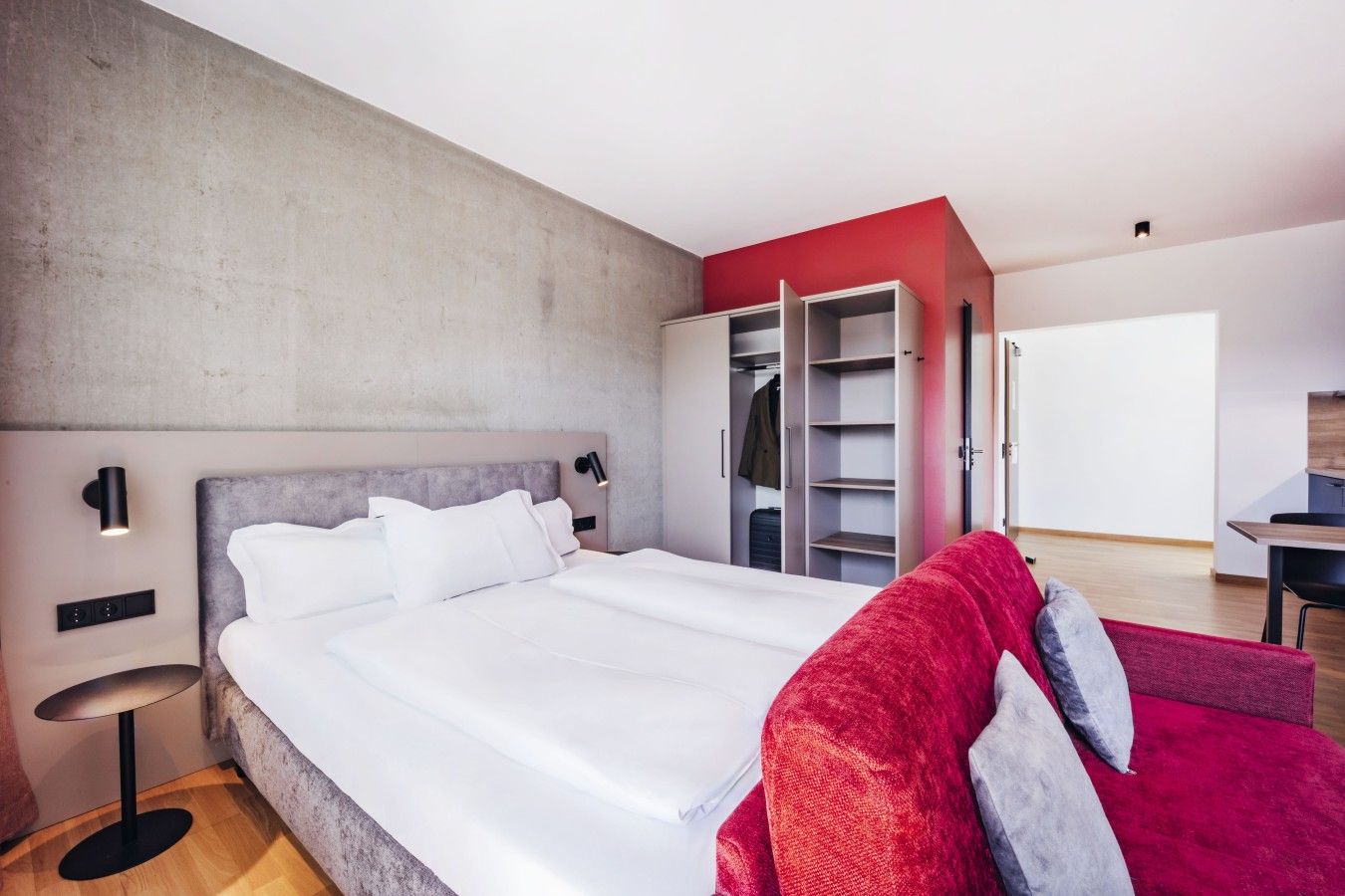 A modern bedroom with a large bed, a wardrobe, a desk, and a red sofa.