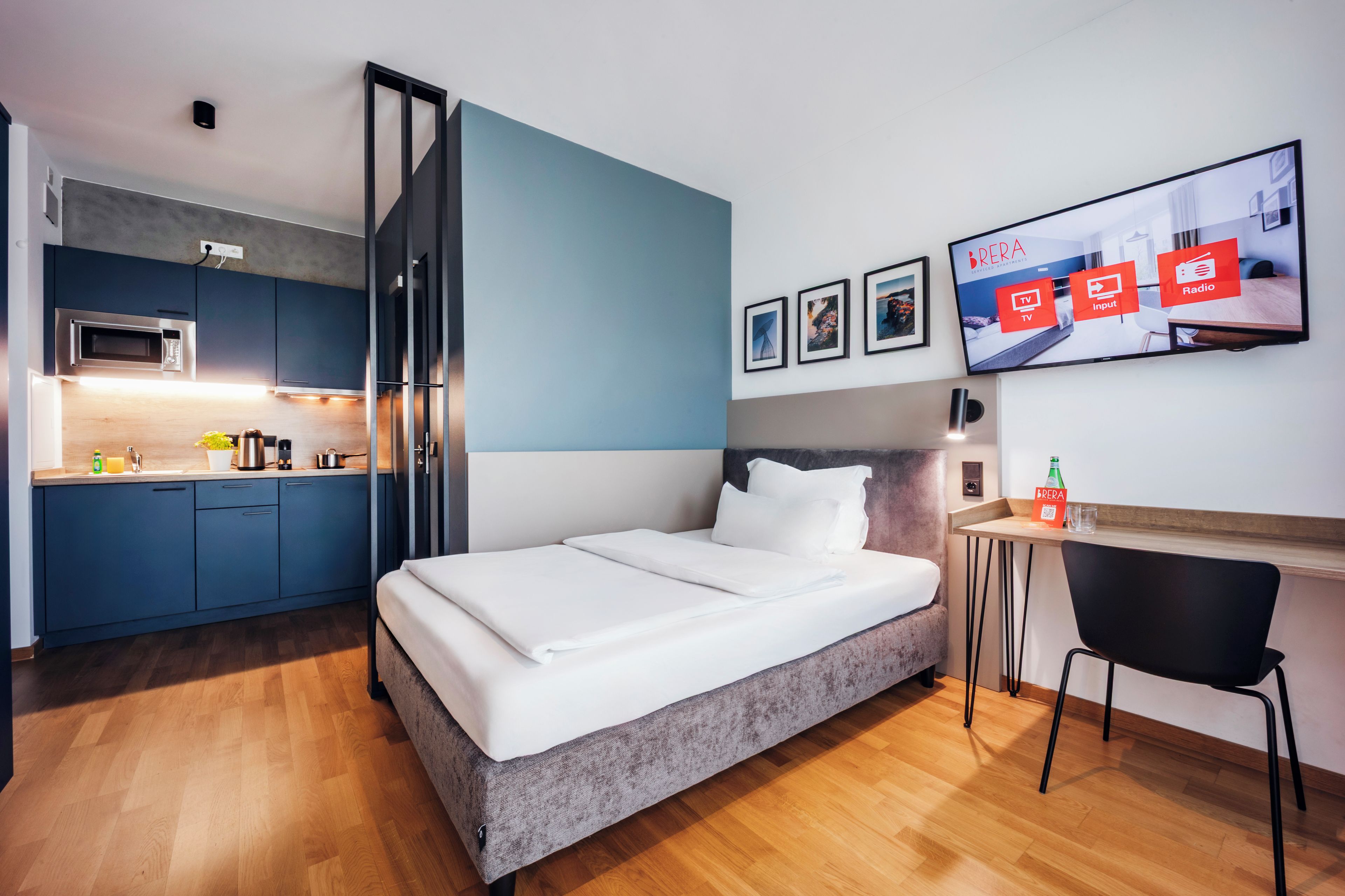 Quarto de hotel compacto com cama, escrivaninha, bancada de cozinha e TV mostrando os serviços do quarto.
