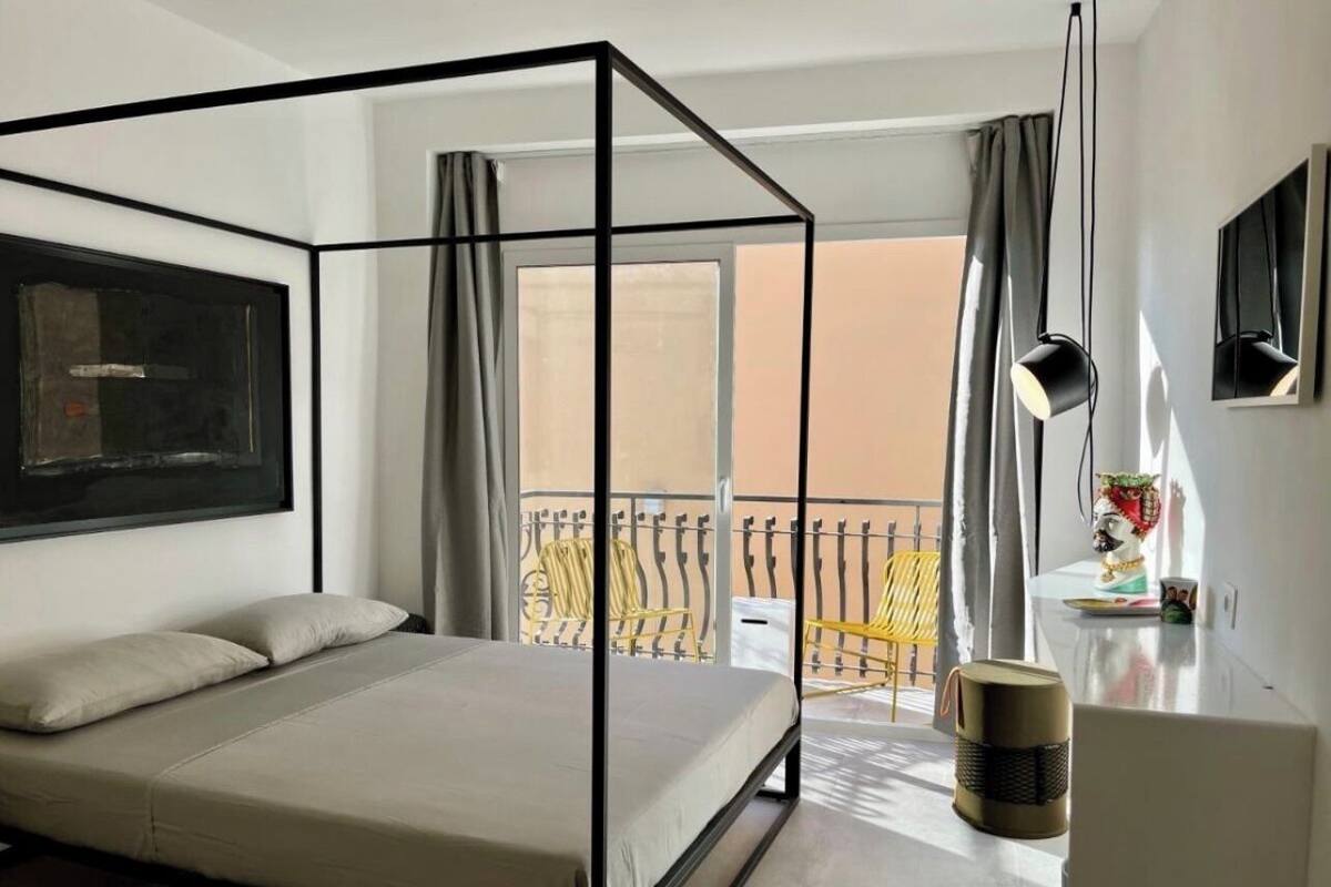 Een slaapkamer met een hemelbed, een balkon met gele stoelen en een groot raam met gordijnen.