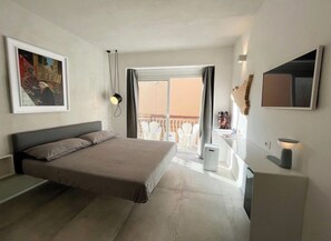 Een moderne hotelkamer met een groot bed, een balkon met witte stoelen, een klein bureau en een wandgemonteerde televisie.