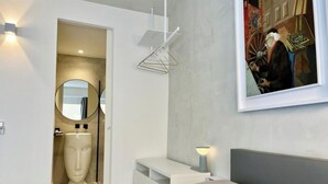 Een moderne slaapkamer met een bed, een nachtkastje, een hanglamp, een schilderij en een badkamer met een spiegel en een bustebeeld.