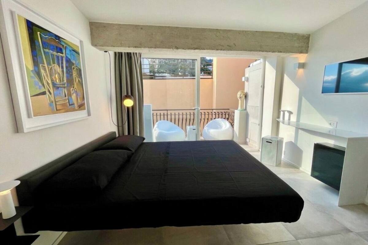 Een moderne hotelkamer met een bed, een schilderij aan de muur, een balkon met stoelen en een aan de muur bevestigde televisie.