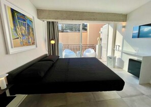 Een moderne hotelkamer met een bed, een schilderij aan de muur, een balkon met stoelen en een aan de muur bevestigde televisie.