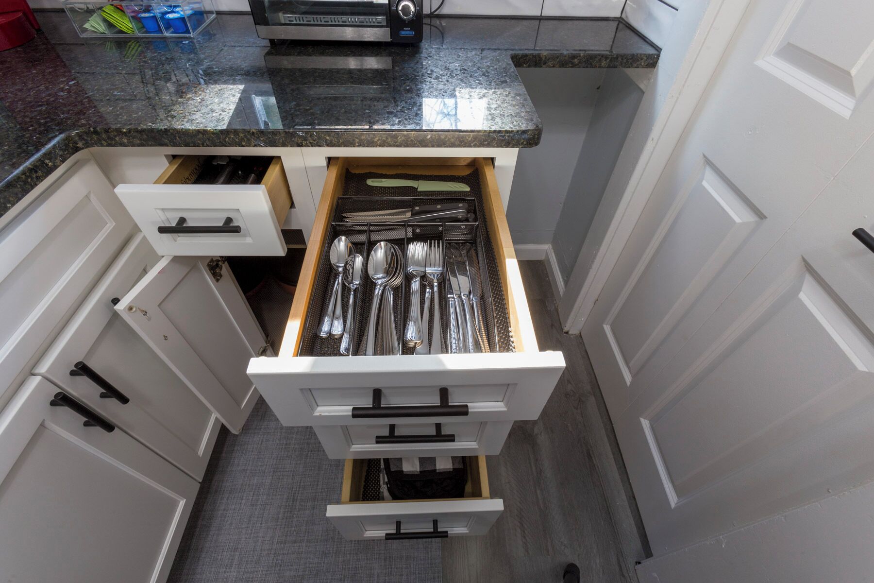 A kitchen with white cabinets, a granite countertop, and a drawer organizer containing silverware.