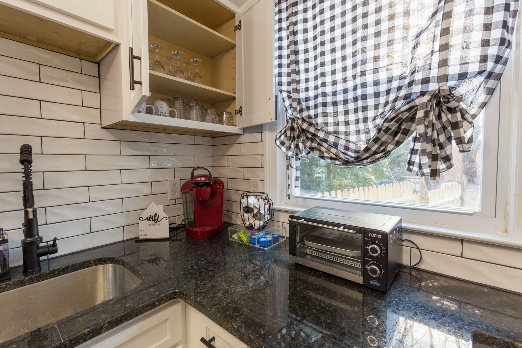Una cocina con armarios blancos, una cafetera roja, un tostador y una ventana con cortinas a cuadros.