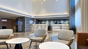 Um saguão de hotel com áreas de estar, um balcão de recepção e um painel de informações digital.