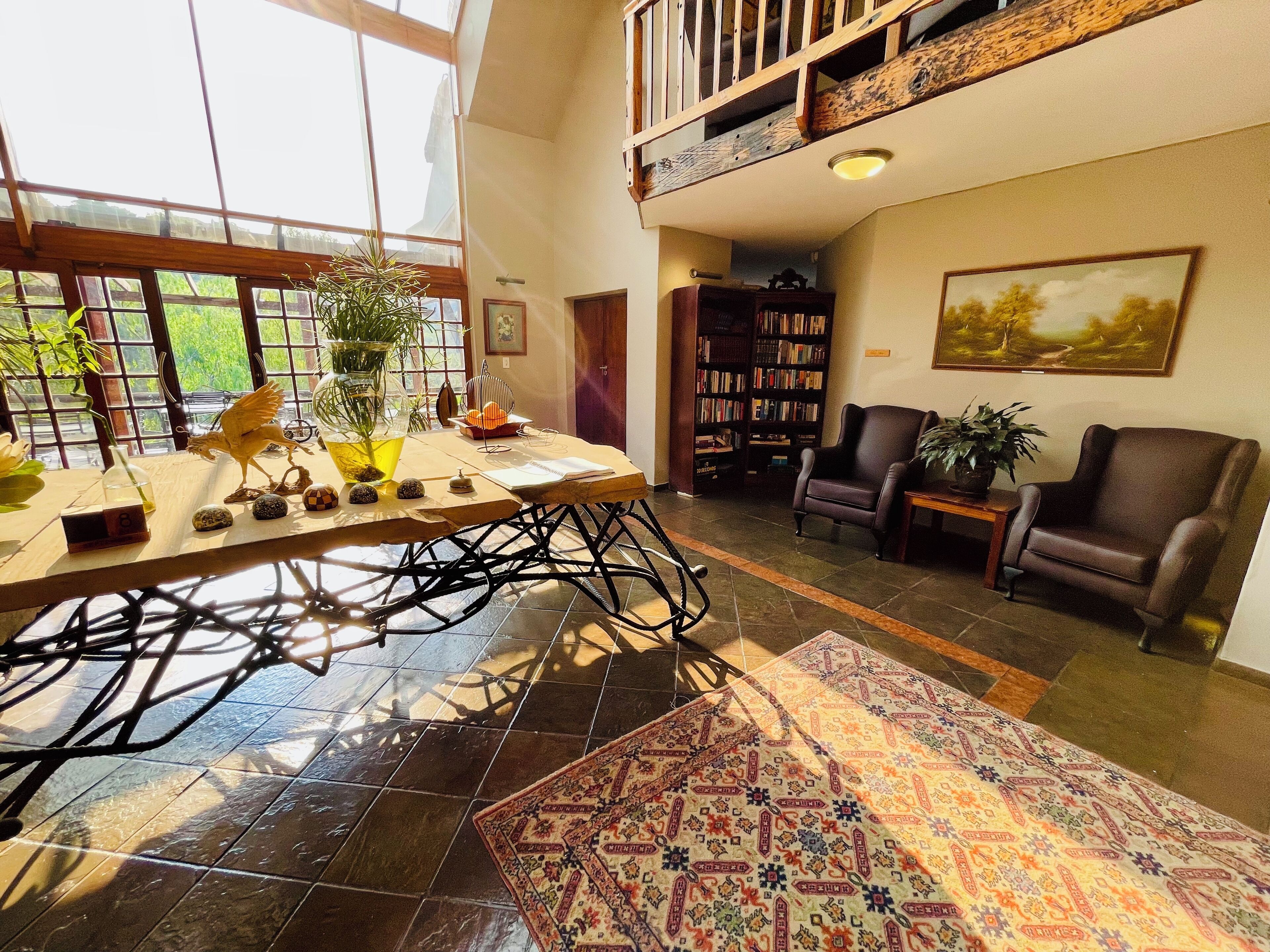 A spacious living area with a high ceiling, a large window, a bookshelf, a dining table, and a patterned rug.