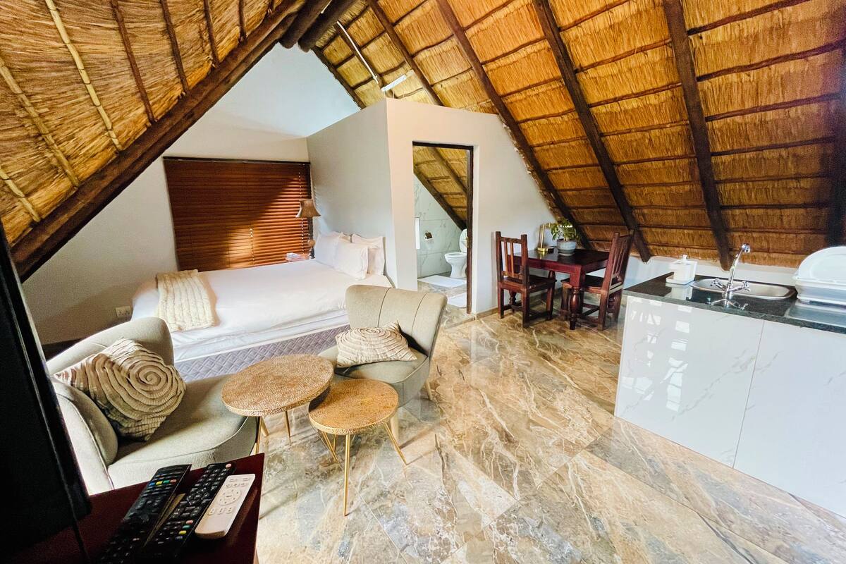 A cozy, compact room with a bed, sofa, and dining area, featuring a thatched roof and a small kitchenette.