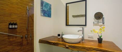 A bathroom with a wooden vanity, a white sink, a mirror, a shower, and a small potted orchid.