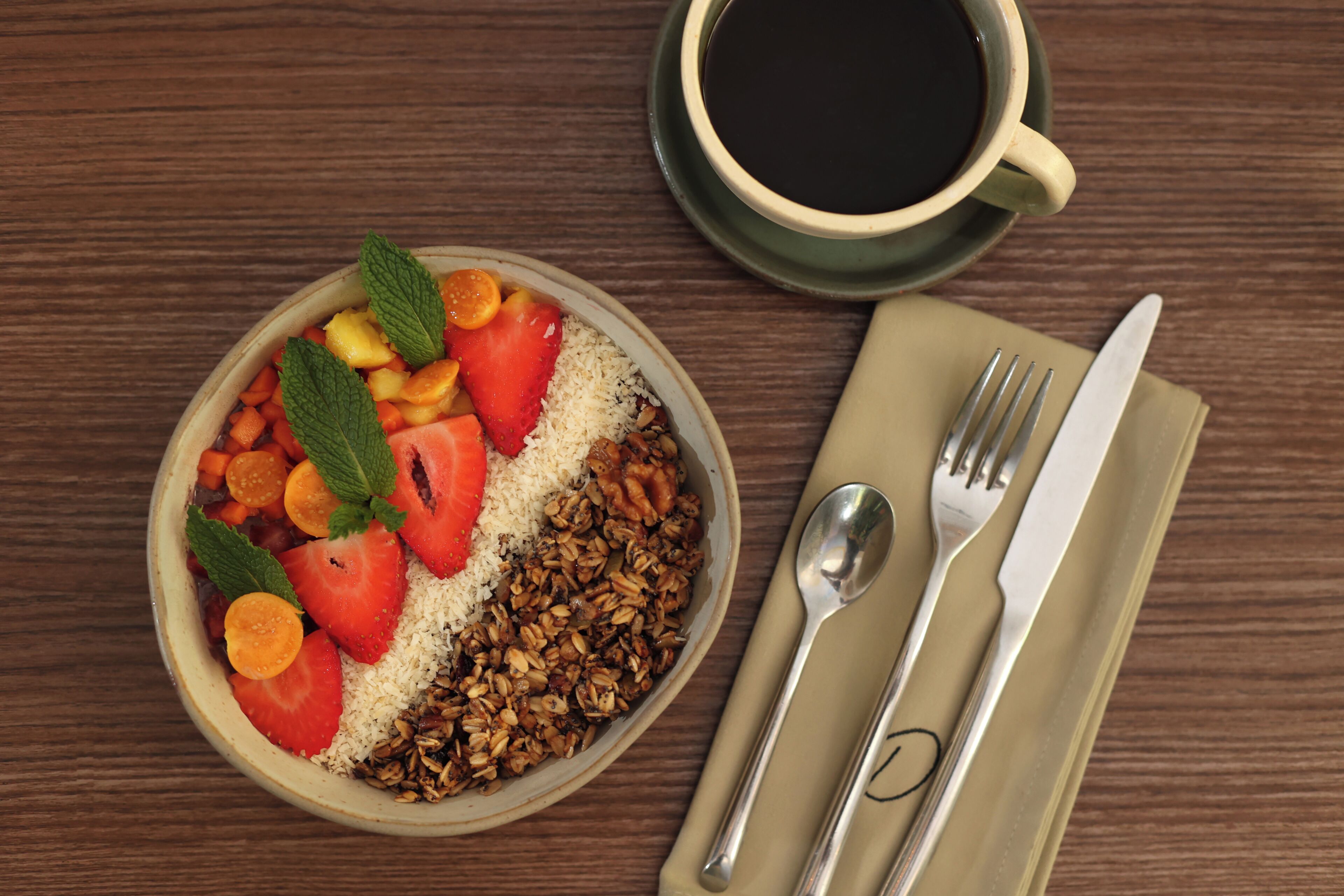 A bowl of cereal with fruits and nuts, a cup of coffee, and cutlery on a wooden table.
