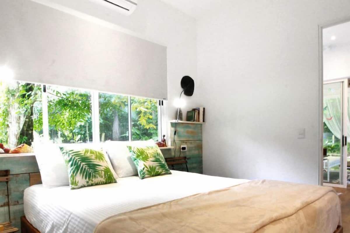 Un dormitorio con una cama grande, una mesita de noche, una lámpara de pared y vistas al exterior con vegetación.