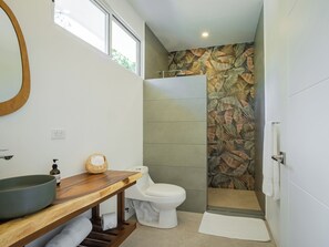 Une salle de bain avec un meuble-lavabo en bois, des toilettes, une douche dont le mur est décoré d’un motif de feuilles, et une fenêtre.