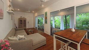 Een moderne woonkamer met een bank, een houten eettafel, plafondventilatoren en grote ramen met uitzicht op groen.