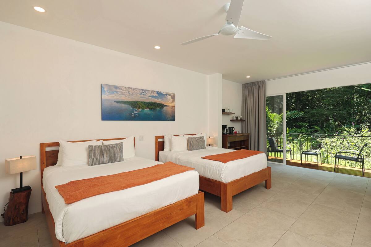 Habitación de hotel con dos camas, ventilador de techo, un cuadro de una escena costera, balcón con sillas y vista a zonas verdes.
