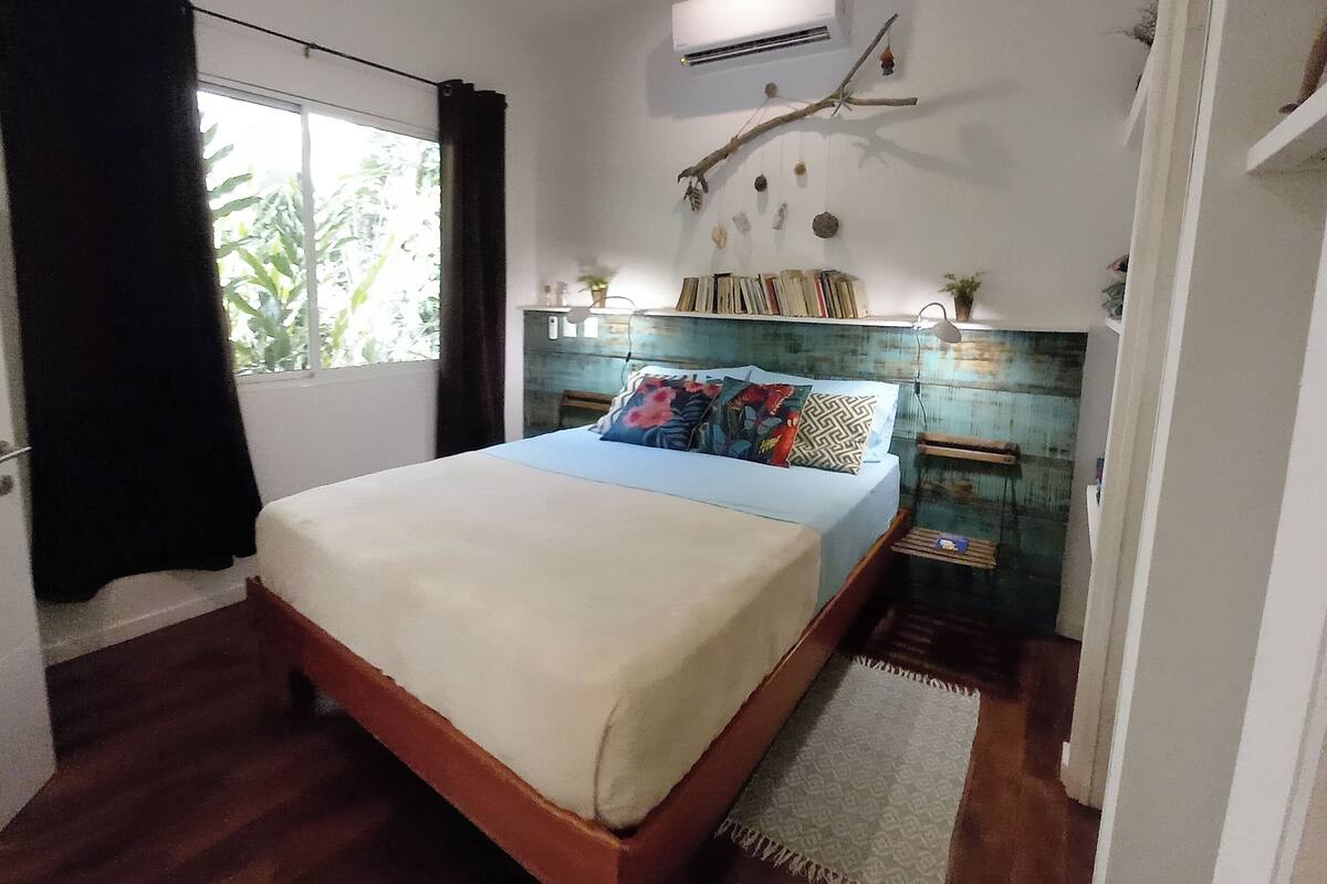 Un dormitorio con cama, ventana con cortinas, aire acondicionado de pared, una rama decorativa y estante con libros y plantas.