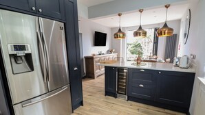 A modern kitchen with a stainless steel refrigerator, a built-in oven, and a wine cooler. There are pendant lights above the island, and a large window provides natural light. - Bryn Gwynedd (Conwy)
