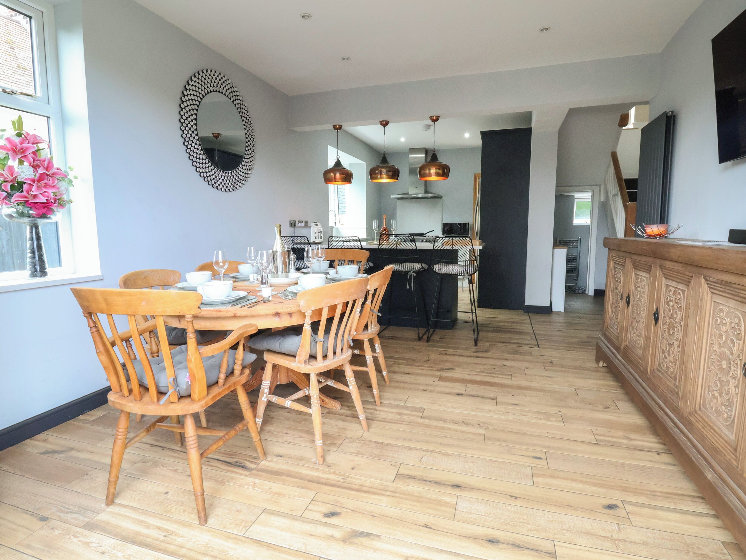 A dining area with a wooden table and chairs, a kitchen in the background, and pendant lighting.